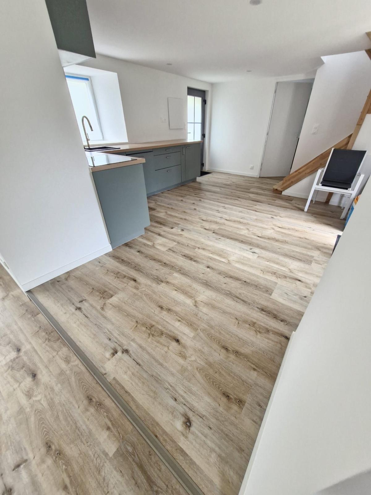 Salle de séjour avec sol en PVC imitation bois clair, cuisine ouverte et escalier en bois.